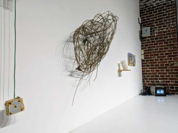 Side-view photograph of an abstract sculpture constructed from willow . The organic structure consists of interconnected, branching forms that weave, cross, and diverge in multiple directions. The piece is suspended against a white wall, creating delicate shadows that emphasize its layered and three-dimensional quality.