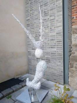 Sculptural work made from upcycled polypropylene strapping and fibreglass rods on a stainless steel frame, resembling a flowing hybrid form inspired by a posy holder interpreted as an oriental fish or dragon-like figure. It stands on a grey pavement slab floor placed in front to a grey bricked wall at teh back and creme painted wall to the side