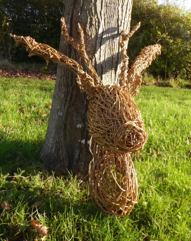 young stag head created from willow resting on grass and against a tree trunk, with bushed in the background